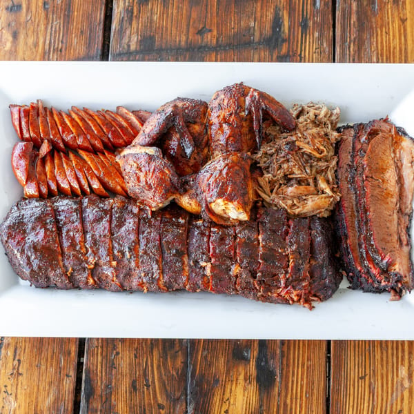A white platter filled with sliced sausage, BBQ chicken, pulled pork, ribs, and brisket.