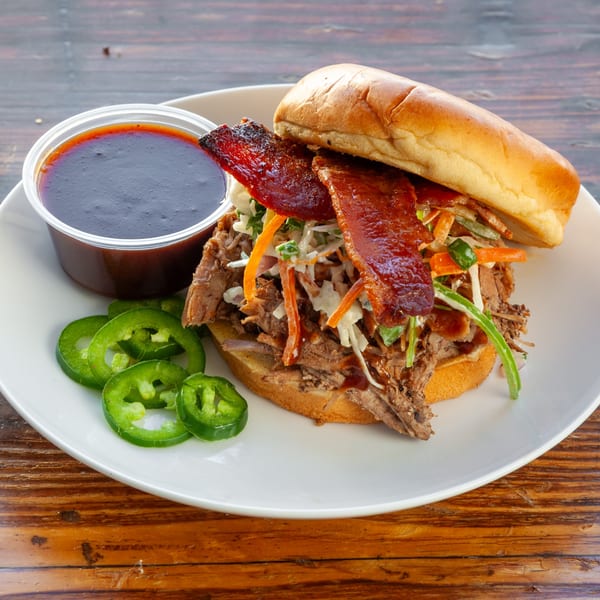 Pulled pork sandwich with bacon, coleslaw, jalapeños, and a side of barbecue sauce on a white plate.