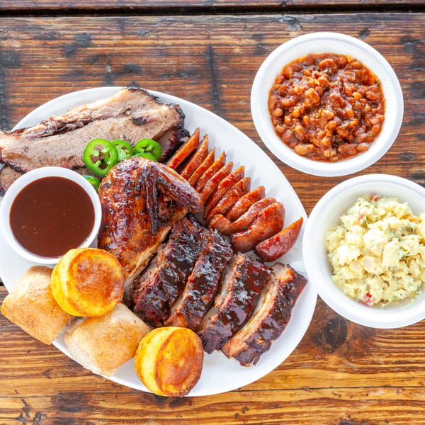 A large platter of barbecue brisket, ribs, chicken, sausage, rolls, sauce, baked beans, and potato salad.